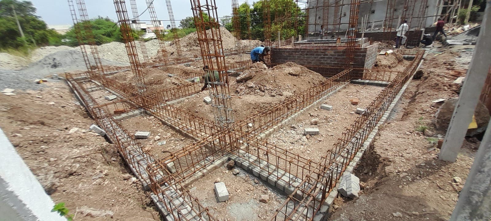 Plinth beam reinforcement laid out on the excavated foundation, with workers and column starter bars